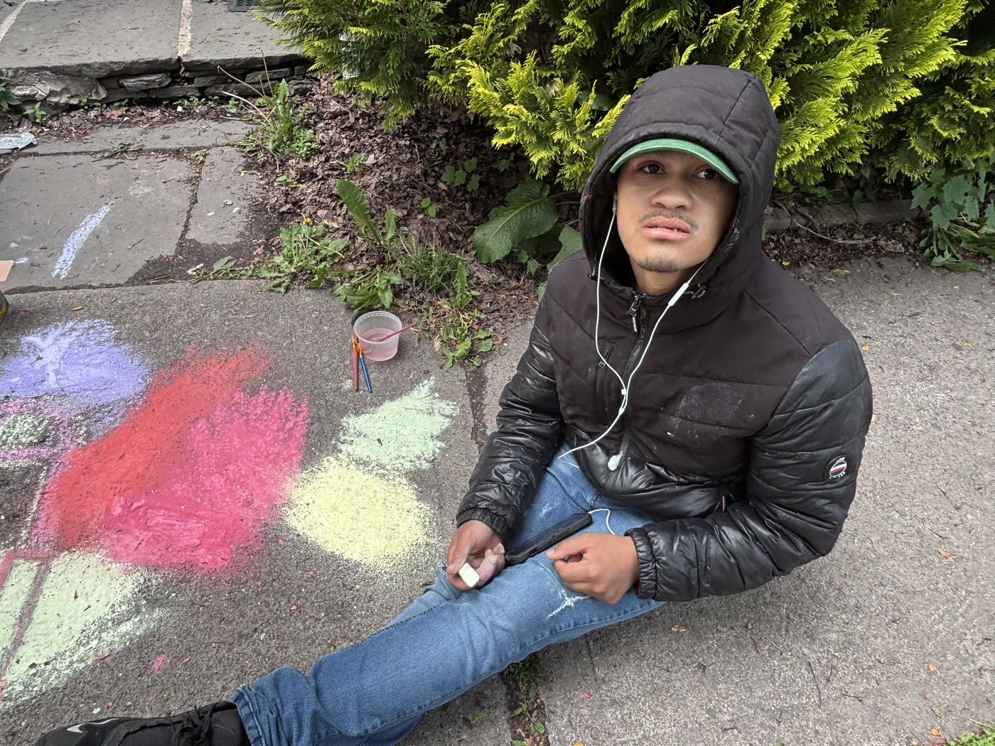 Bashir sitting on the sidewalk beside bright chalk flowers he drew.